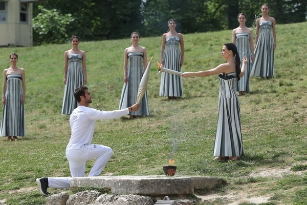 扮演最高女祭司的演員瑪麗·米娜（前右）點(diǎn)燃第一棒火炬手——希臘運(yùn)動(dòng)員、東京奧運(yùn)會(huì)男子賽艇項(xiàng)目金牌得主斯特凡諾斯·恩圖斯科斯手中的火炬。新華社記者 趙丁喆 攝